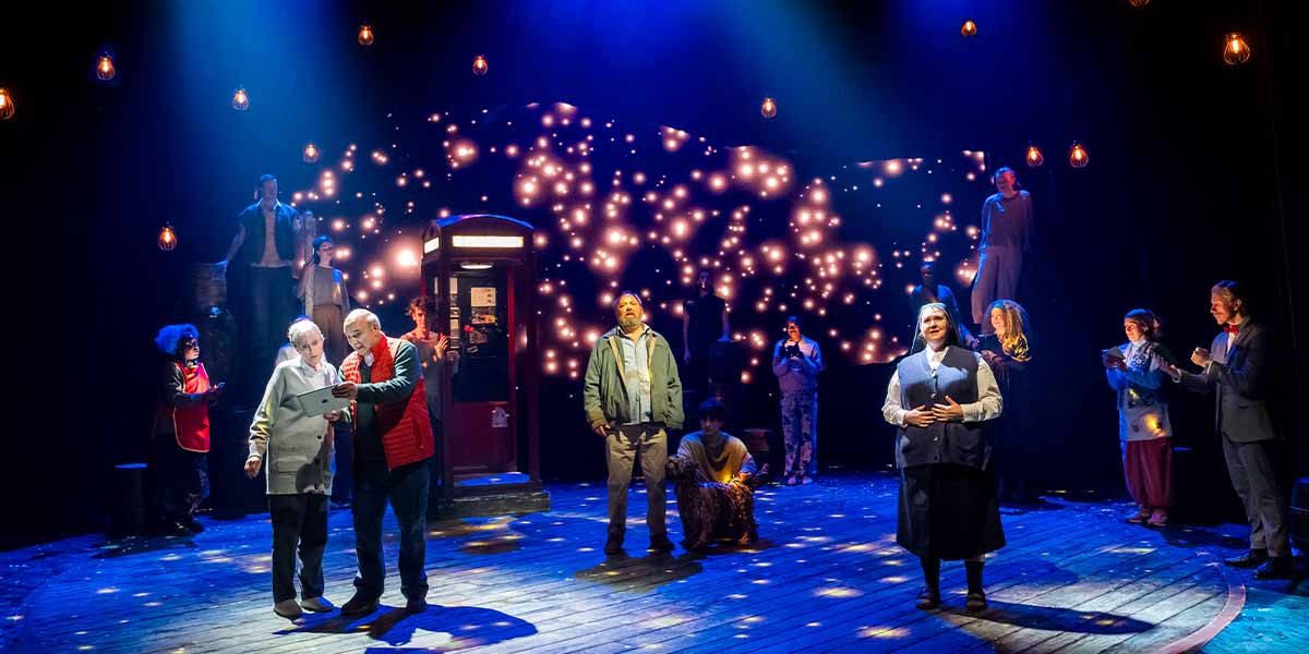 Jenna Russell and Peter Polycarpou interact centre stage, surrounded by cast members against a starry backdrop.