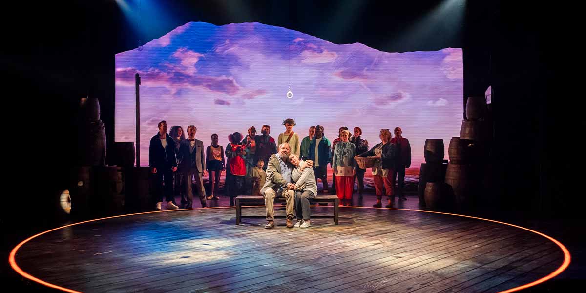 Mark Addy and Jenna Russell seated on a bench, surrounded by a cast against a scenic backdrop.
