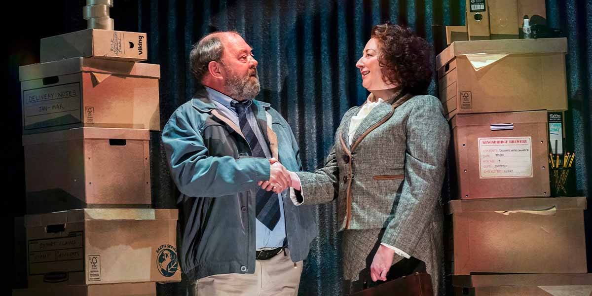 Mark Addy and a woman shake hands in front of stacked cardboard boxes, smiling warmly.