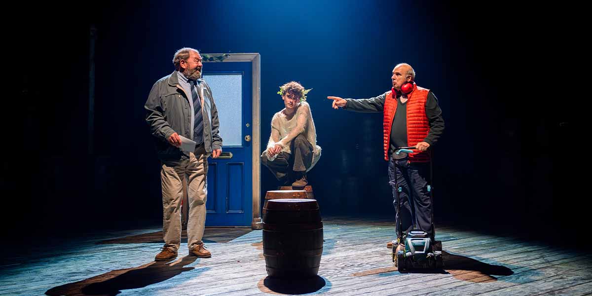 Three male actors on stage: one crouches on a barrel, while the others engage in conversation.
