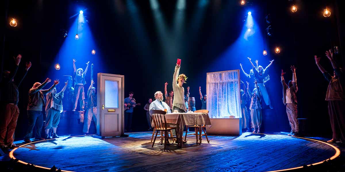 Mark Addy and Noah Mullins perform on stage, surrounded by cast members raising their arms in celebration.