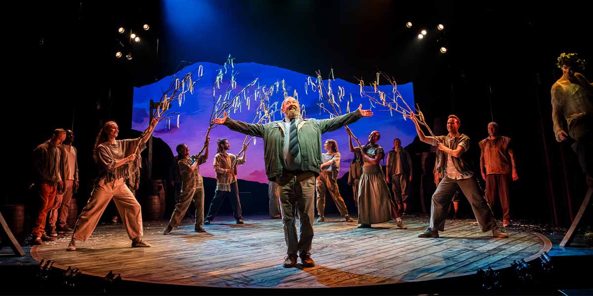 Mark Addy as Harold Fry stands centre stage, surrounded by cast members in vibrant costumes, with dramatic lighting.