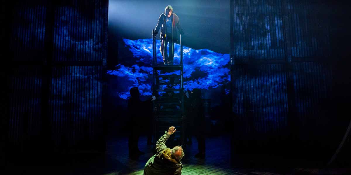 Noah Mullins stands on a ladder, reaching down to an outstretched hand in a dramatic, dimly lit scene.