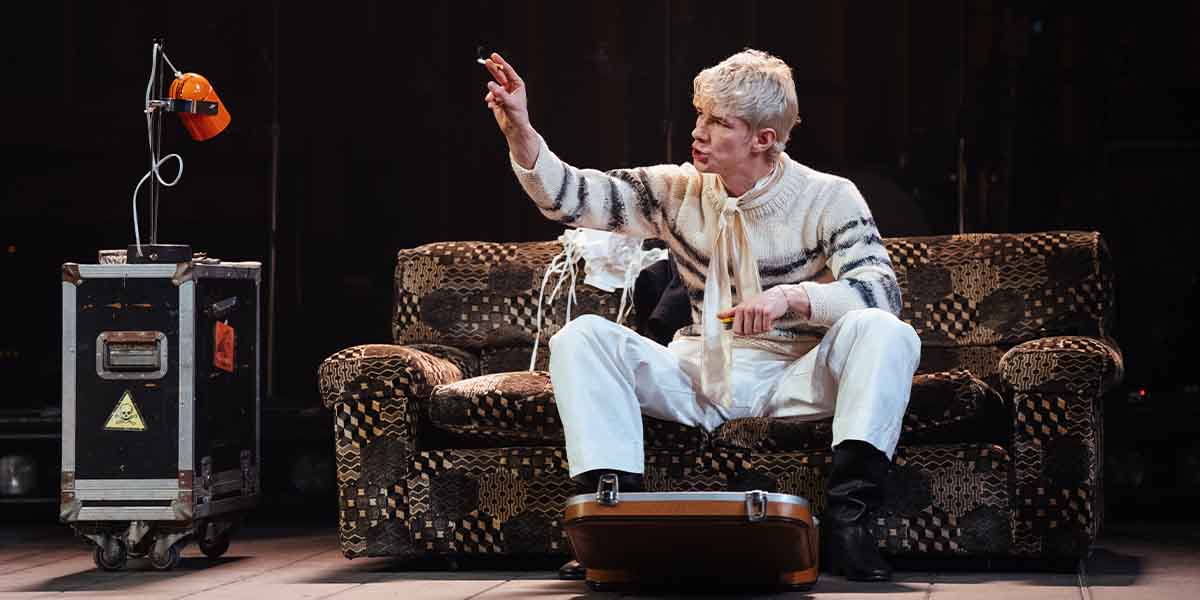 Samuel Jordan sits on a patterned sofa, gesturing animatedly while holding a cigarette and yellow object.