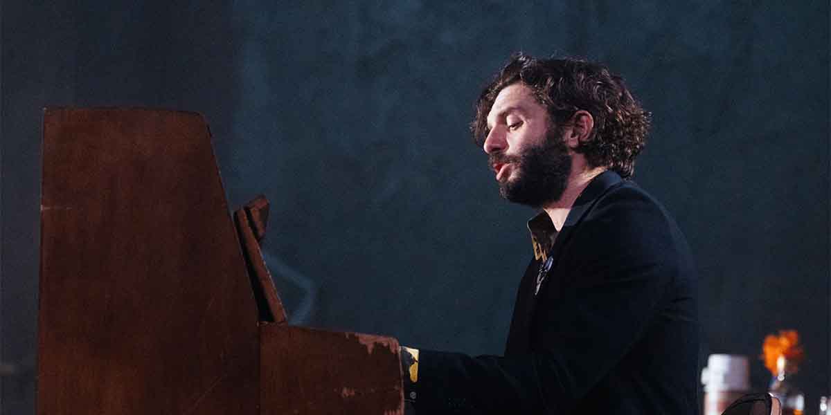 Michael Fox, dressed in black, plays a wooden piano, with a blurred background and warm lighting.