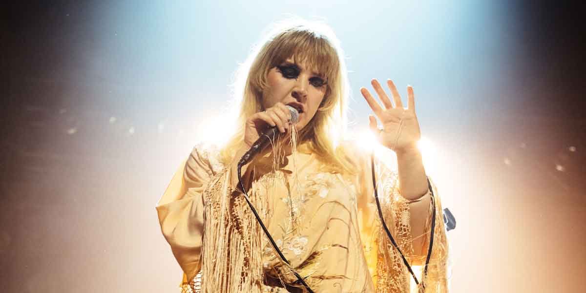 Rebecca Lucy Taylor performs on stage, wearing a golden outfit, with dramatic lighting illuminating her.