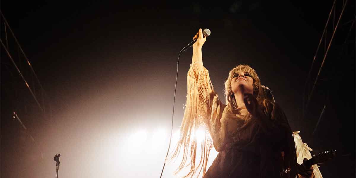 Rebecca Lucy Taylor, wearing a flowing outfit, passionately sings into a microphone against a dramatic, illuminated background.