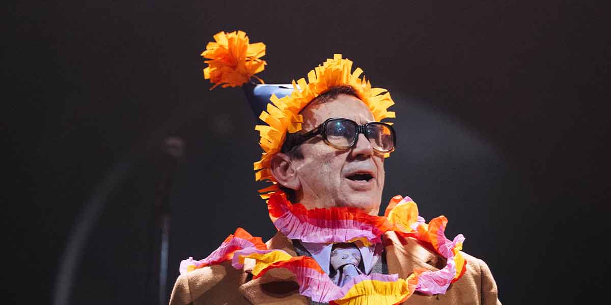 Phil Daniels performs in a colourful costume with a party hat and floral decorations against a dark background.