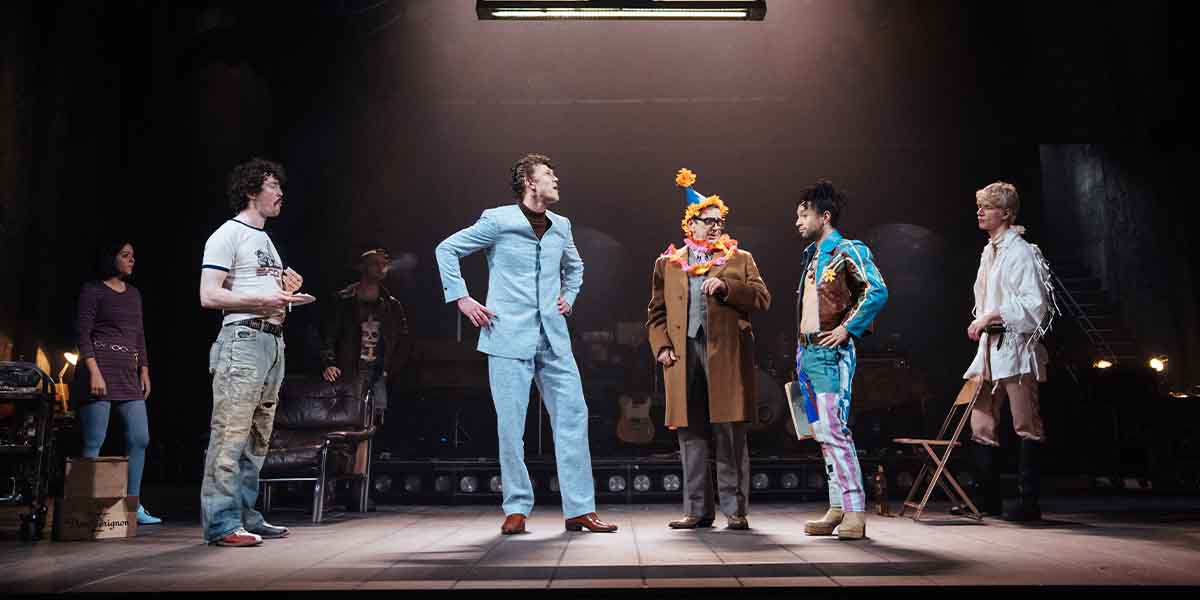 Six actors perform on stage, wearing colourful costumes, with dramatic lighting and a dark background.