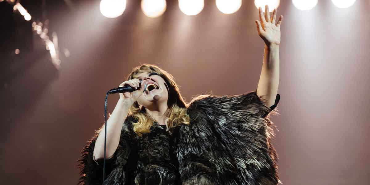 Rebecca Lucy Taylor performs passionately on stage, wearing a fur coat, with bright stage lights above.