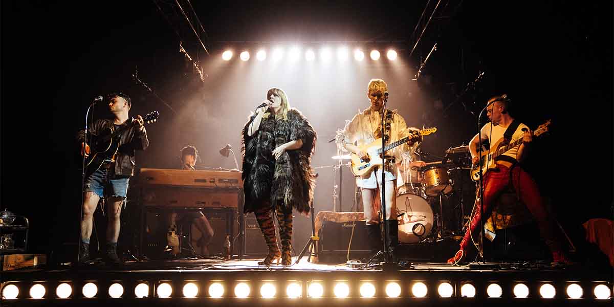 Noah Weatherby, Rebecca Lucy Taylor, Samuel Jordan, and Jojo Macari perform on stage with instruments and dramatic lighting.