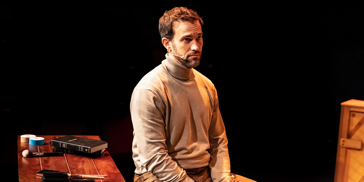 A person in a beige turtleneck sits pensively on a stage, surrounded by props and a dark background.