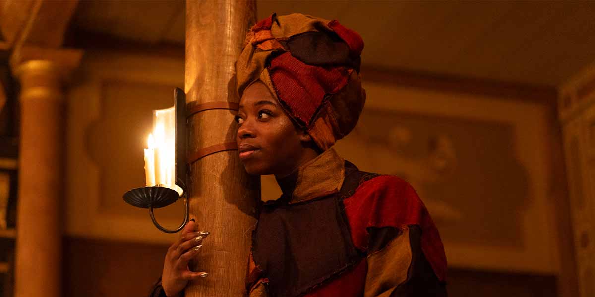 Aminita Francis as SK Good leans against a wooden pillar, illuminated by a candle, wearing a colourful costume.