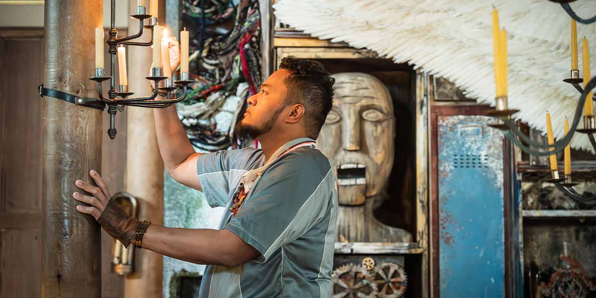 Faizal Abdullah as Caliban adjusts a candle on a chandelier, surrounded by an eclectic, textured backdrop.