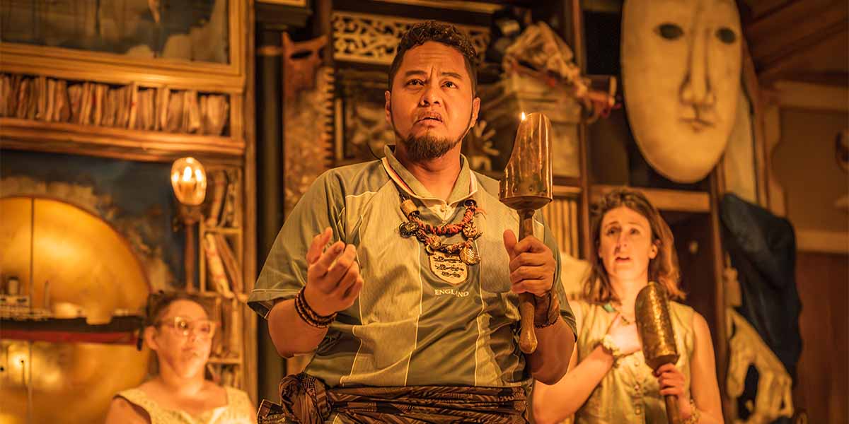A performer holds a bell and gestures expressively, flanked by two women in a theatrical setting.