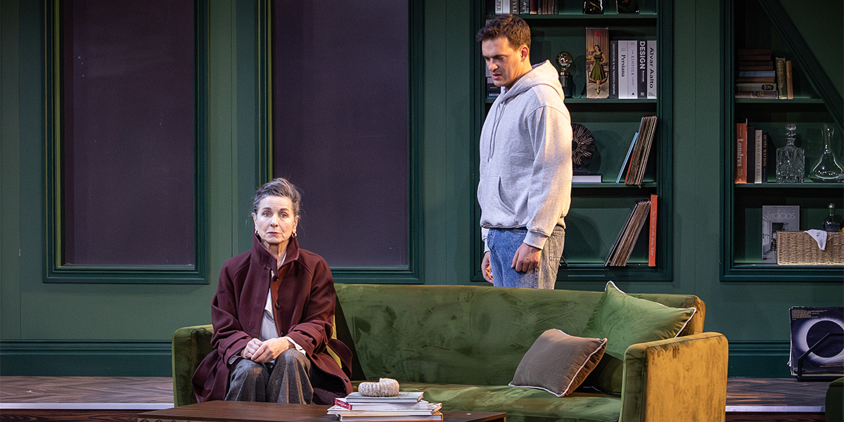 One person sits on a green sofa. Another person wearing a grey hoodie stands behind them.