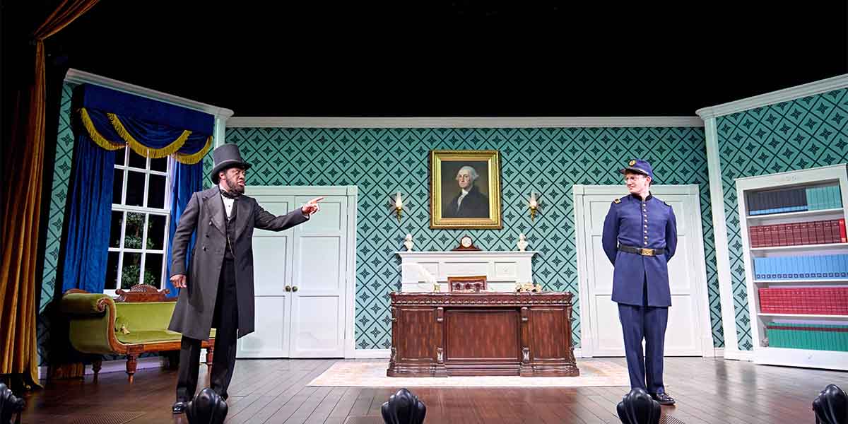 A man in a top hat gestures, while another in a uniform stands by a desk in a theatre.
