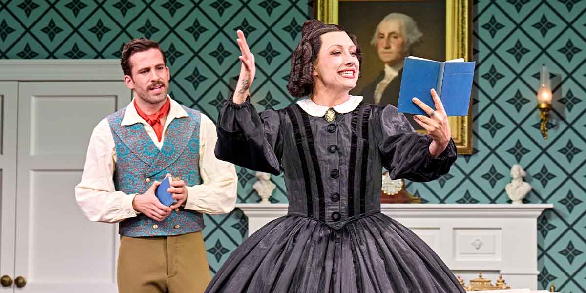 A woman in a black dress gestures while reading from a blue book; a man watches her.
