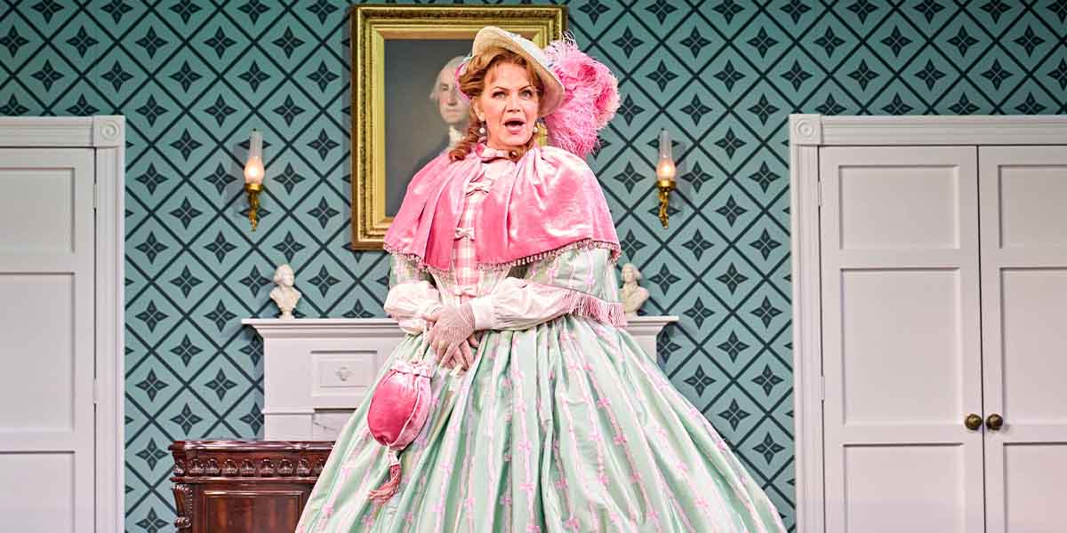 A woman in a pink and green Victorian dress stands dramatically, with a patterned wallpaper and furniture behind her.