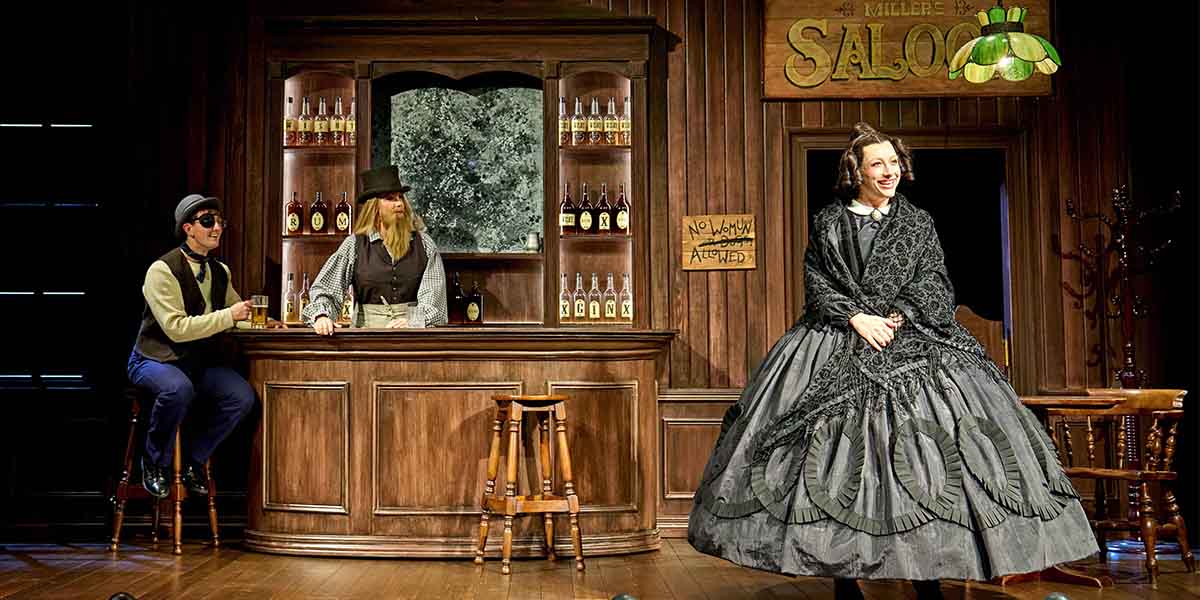A woman in a large black dress stands in front of a bar with two men in period costumes.