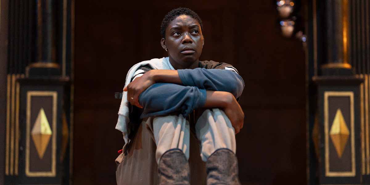 Selina Jones as Azure sits on stage, hugging their knees, with a contemplative expression.