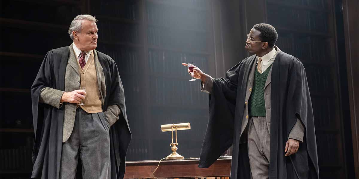 Two men in formal attire stand in a library setting; one holds a drink, gesturing.