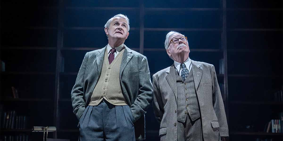 Two men in vintage suits look upwards, surrounded by a dimly lit library backdrop with shelves.