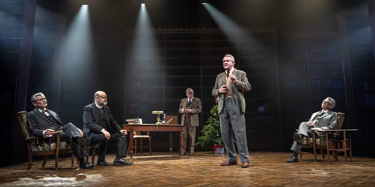 Five men in formal attire engage in conversation on stage, with a desk and bookshelves in the background.
