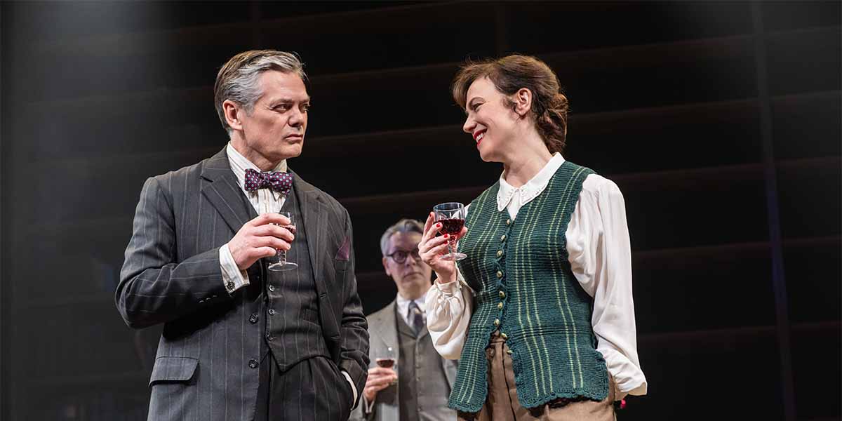 Two elegantly dressed characters hold glasses while smiling at each other, with a third figure in the background.