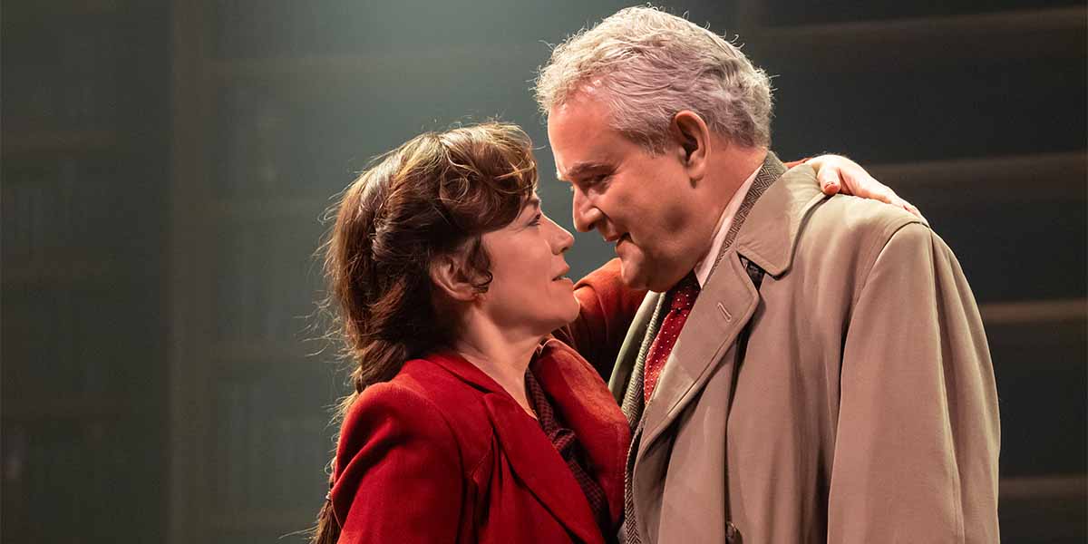 A woman in a red jacket and a man in a beige coat share an intimate moment on stage.