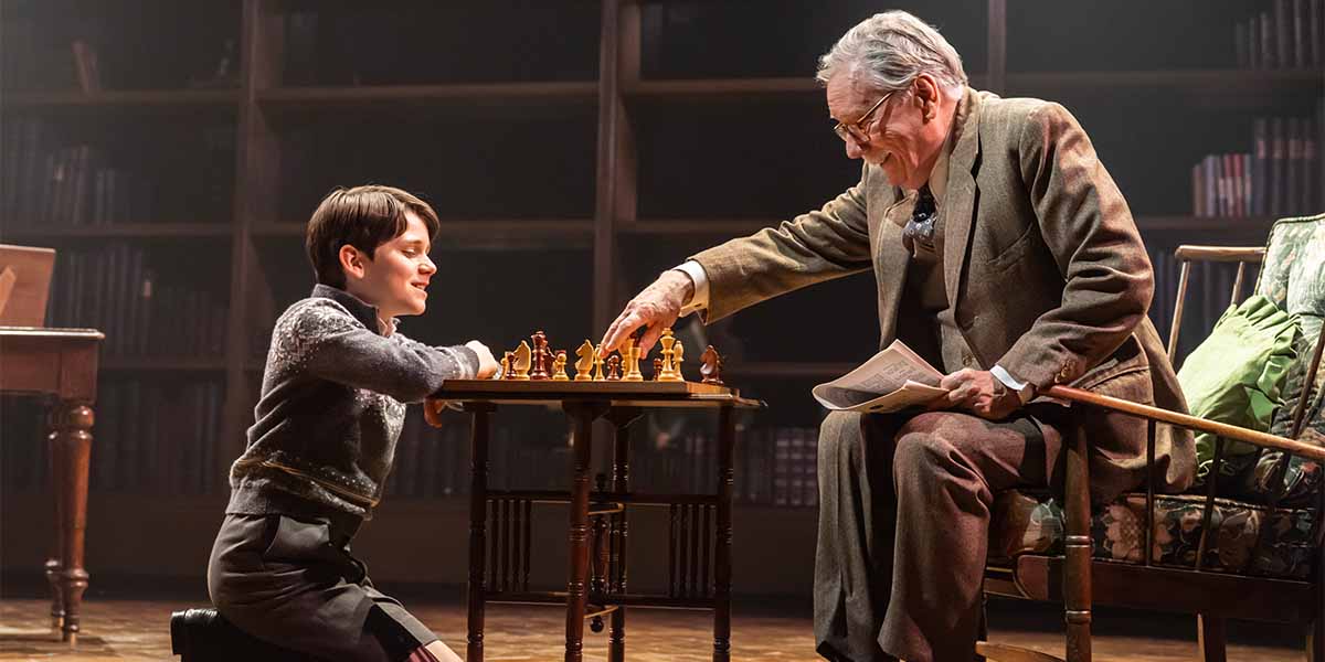 A young boy and an elderly man play chess at a table, surrounded by bookshelves in a warm setting.