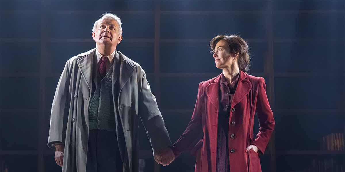Two actors stand on stage, holding hands, looking upwards; one wears a grey coat, the other a red coat.