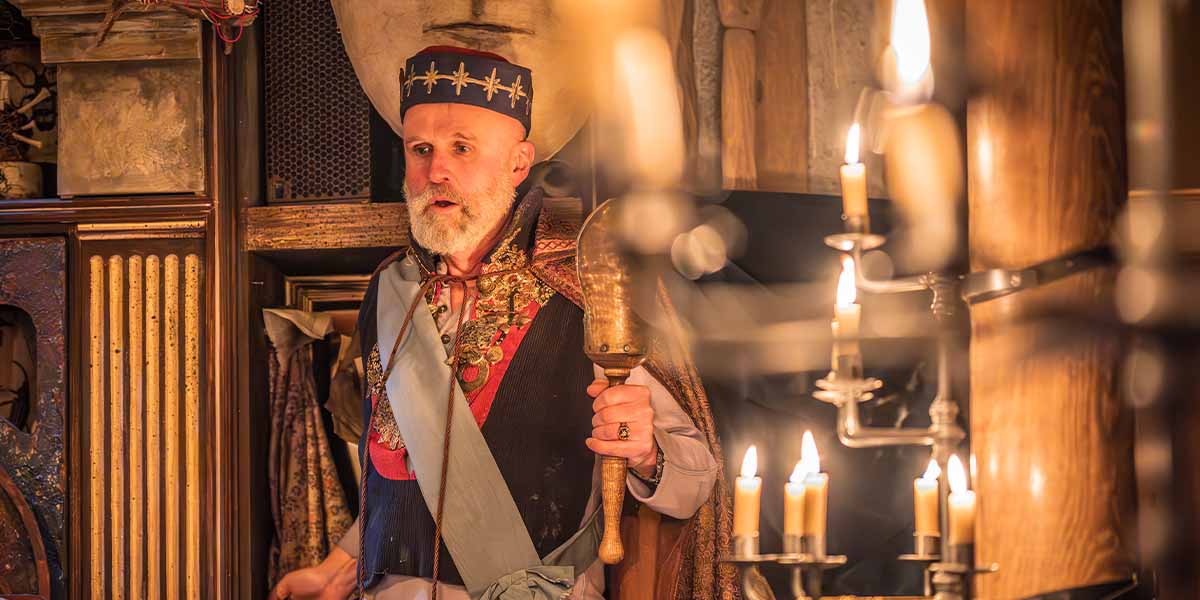 Tim Crouch as Prospero, holding a staff, with candles and ornate background elements in dim lighting.