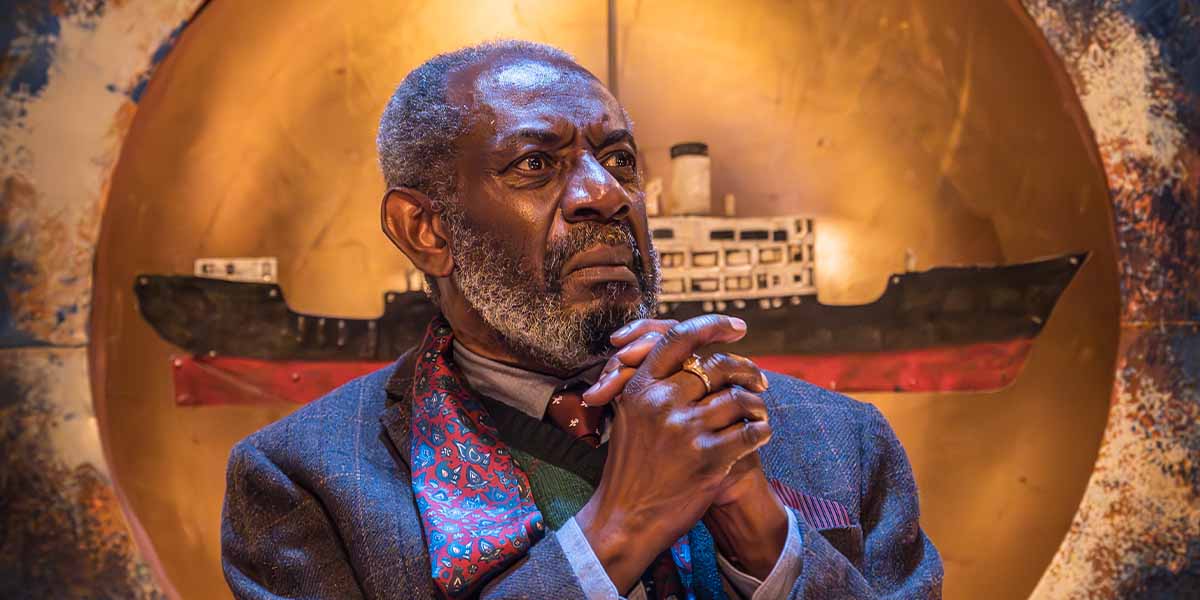 Tyrone Huggins as Gonzala, looking contemplative with clasped hands, against a circular, ship-themed backdrop.