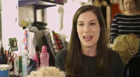 A woman with short brown hair smiles while holding a theatre script, surrounded by colourful stage props.
