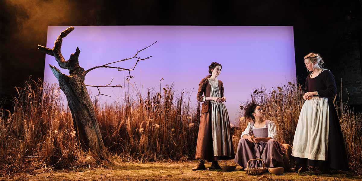 Two people are on stage surround by tall grass, one is standing the other is sitting on the floor.