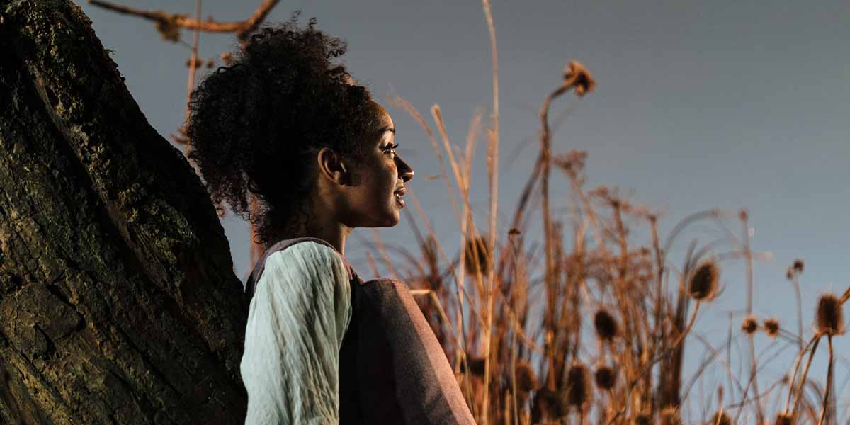 A person with curly hair leans against a textured tree trunk, surrounded by tall, dry grasses.
