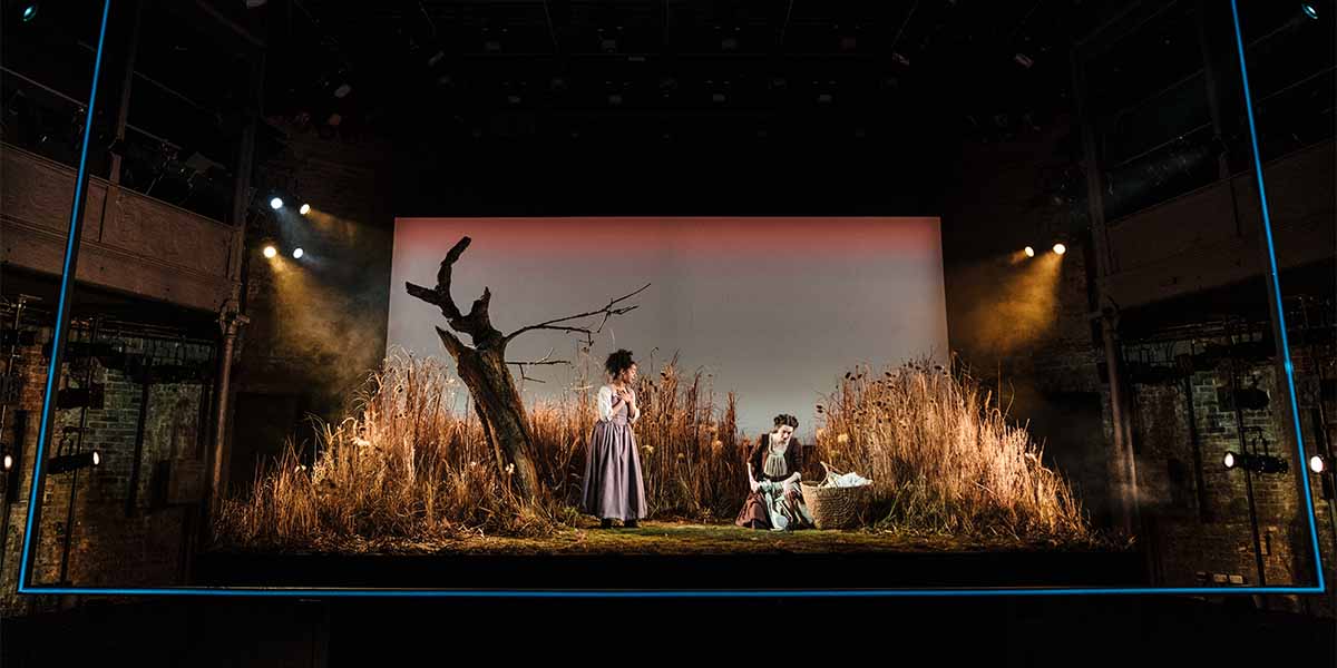 Two people in period costumes stand on a stage with tall grass and a bare tree, dramatic lighting.