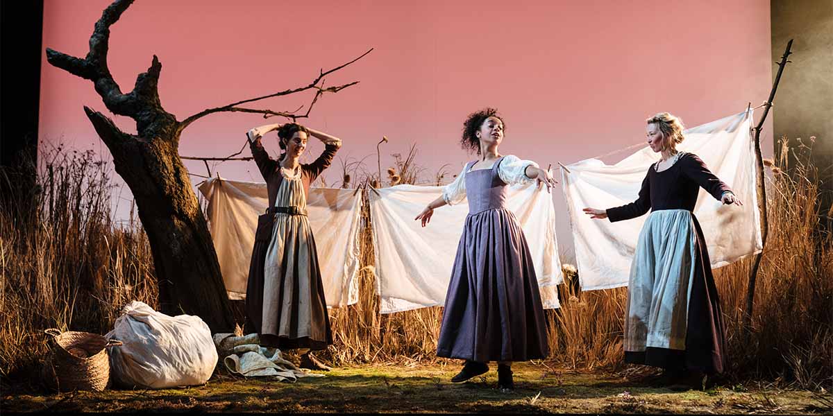 Two people in period clothing stand in front of a washing line covered in white sheets.