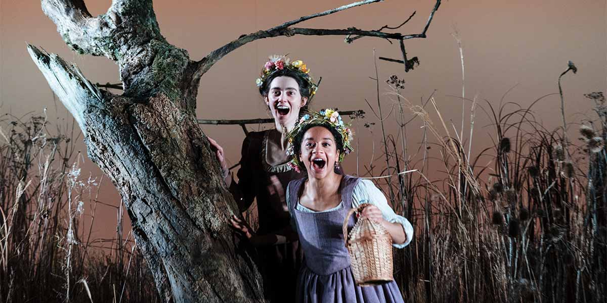 Two people in period clothing stand in front of a tree laughing.