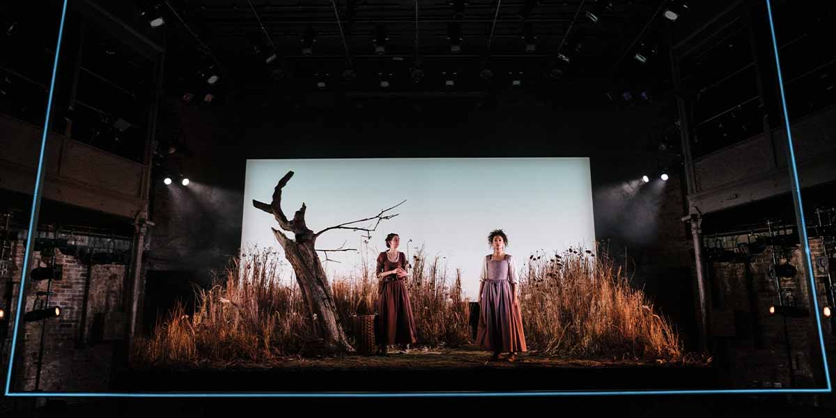 Two women in period costumes stand on stage, surrounded by tall grasses and a bare tree.