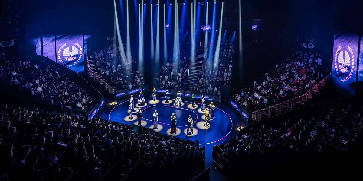 A group of actors in elaborate costumes perform on stage, with dramatic lighting and a dark backdrop.