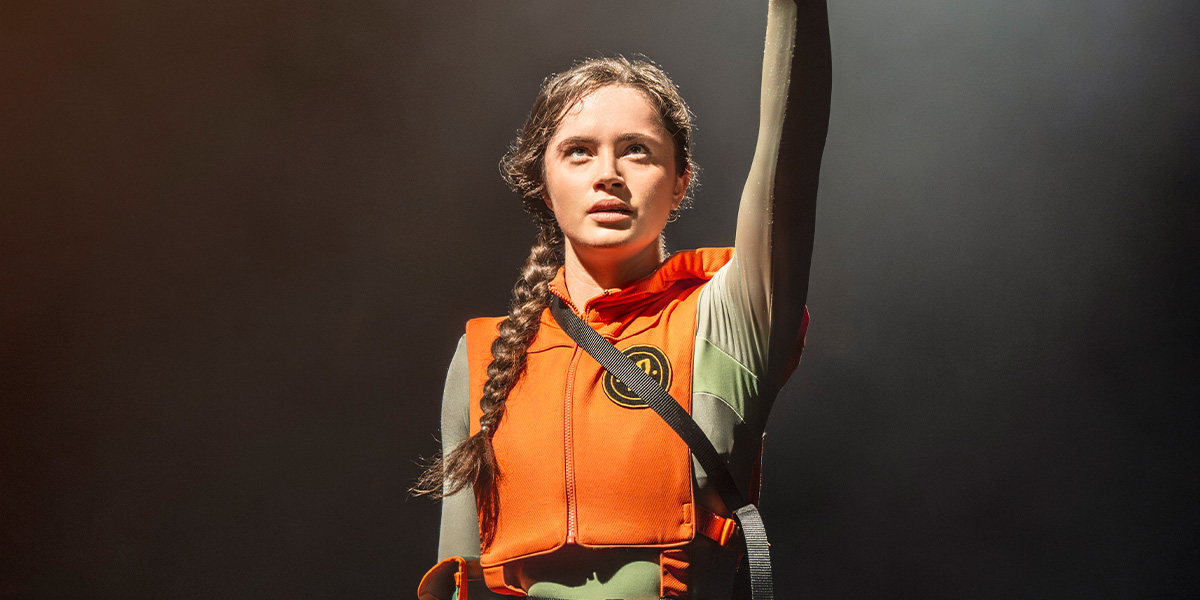 Mia Carragher as Katniss Everdeen raises her arm, wearing an orange life vest against a dark background.