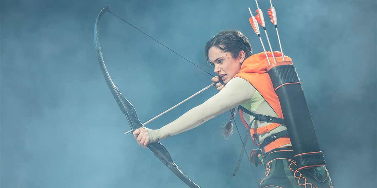 Mia Carragher as Katniss Everdeen, aiming an arrow with a bow, dressed in an orange and black outfit.