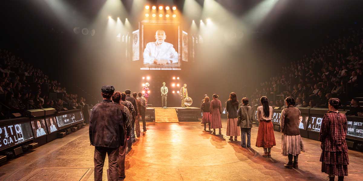 The cast of The Hunger Games on stage, dressed in elaborate costumes, performing in a dramatic setting.