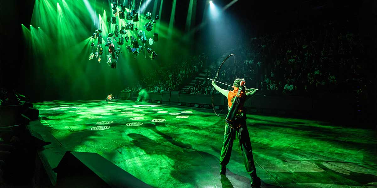 Mia Carragher as Katniss Everdeen stands confidently, bow drawn, in a dramatic stage setting with dark textures.