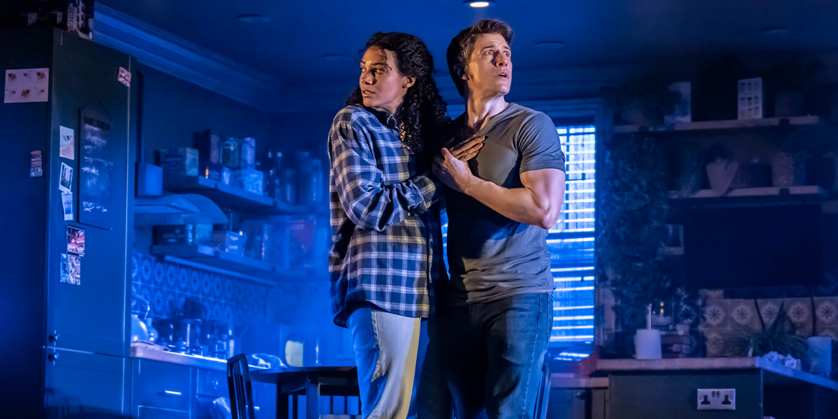 two people stand in a dimly lit room, looking intently at a flickering light source.