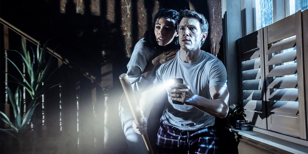 two people stand in a dimly lit room, looking intently at a flickering light source.