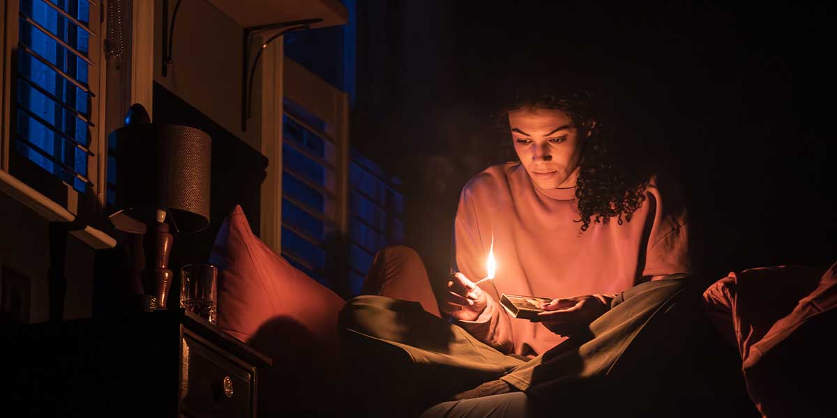 A young woman sits cross-legged on a bed, holding a lit candle and looking intently at it.