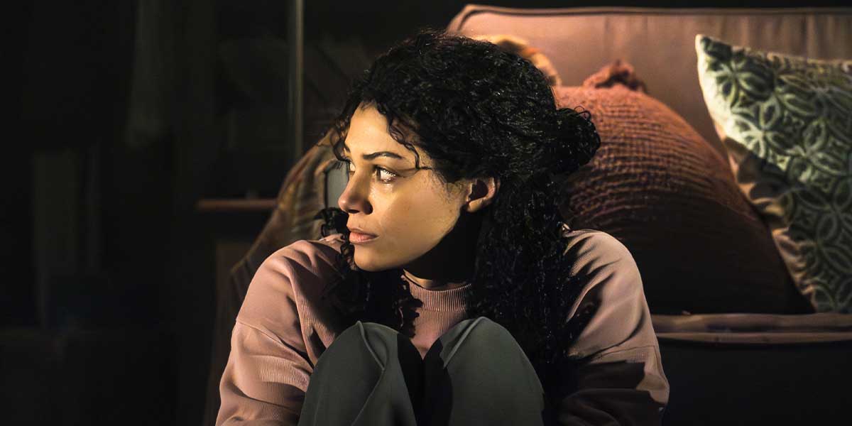 A woman with curly hair sits on the floor, looking anxiously towards the side, surrounded by cushions.